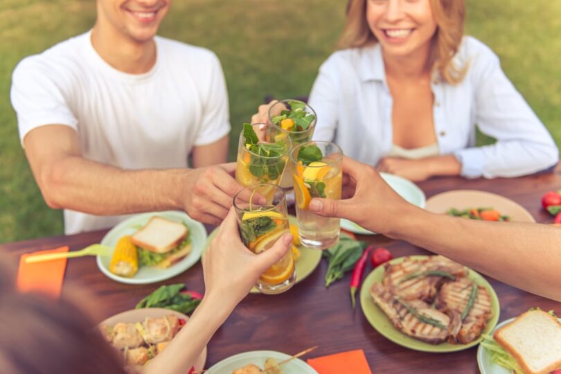 Friends toasting with drinks at an outdoor picnic table with food plates under sunny skies.