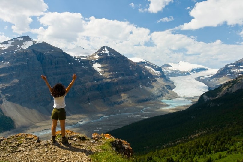 Person standing on a cliff with arms raised, overlooking a valley with mountains and glaciers under a partly cloudy sky.