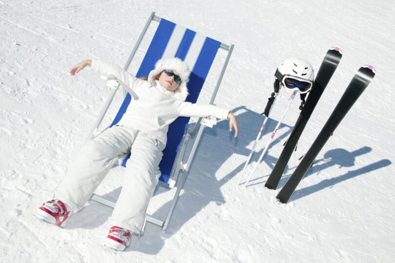 Person in white outfit relaxing on a lounge chair in the snow with skis and poles nearby.