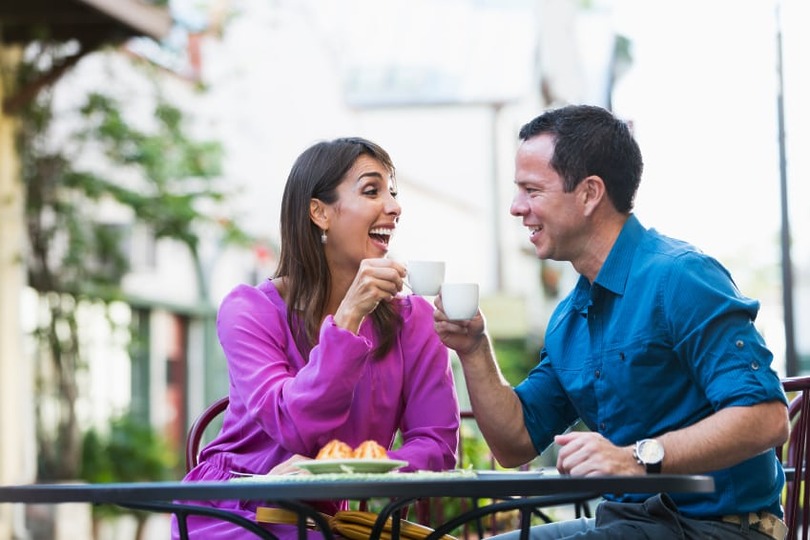 A woman and man laughing while clinking coffee mugs at an outdoor café table, with pastries in front of them.