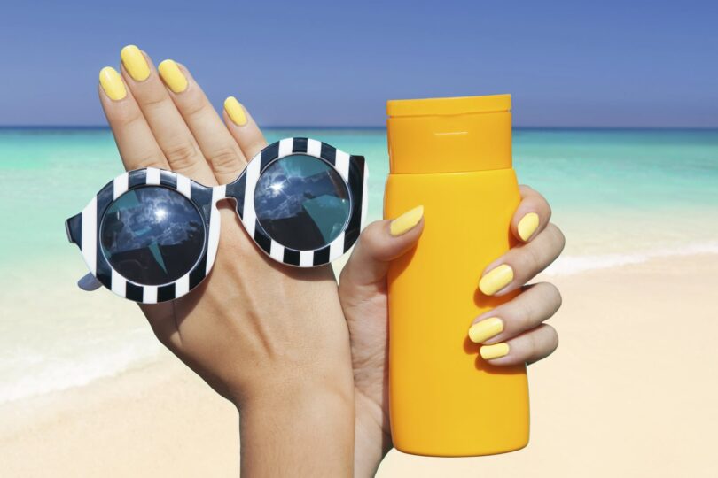 Person holding orange sunscreen bottle and sunglasses with yellow nails, beach and ocean in the background.