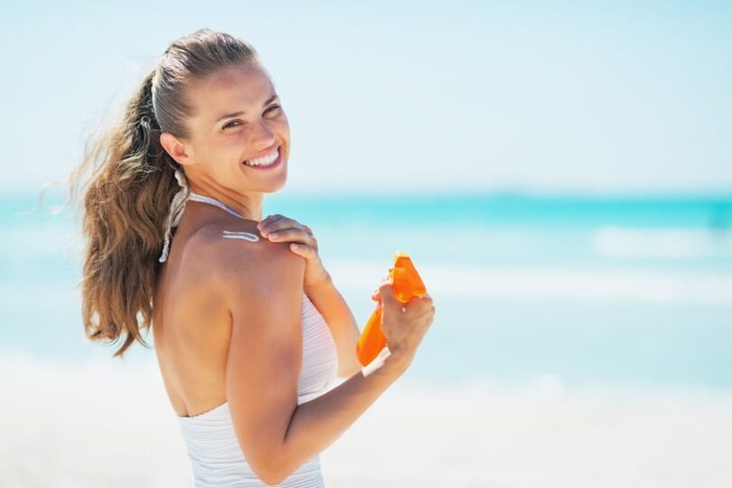 Woman smiling at beach applying sunscreen on her shoulder, clear blue sky and ocean in the background.