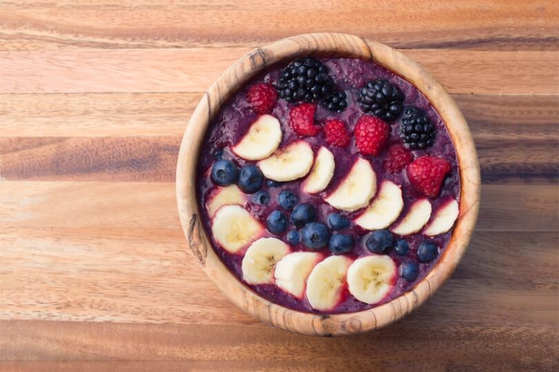 Bowl of acai smoothie topped with banana slices, blueberries, raspberries, and blackberries on a wooden table.