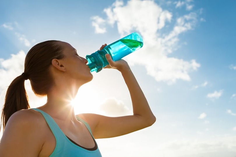 Woman in athletic wear drinks from a blue water bottle under a sunny sky with clouds.