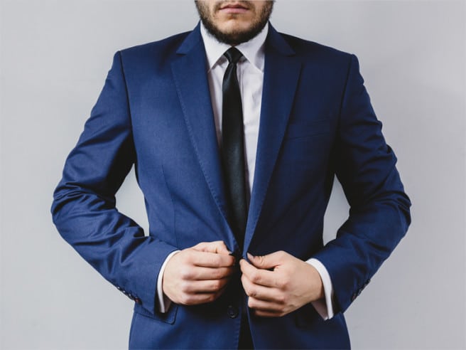 Man in blue suit adjusting jacket with hands, wearing a black tie against a plain background.