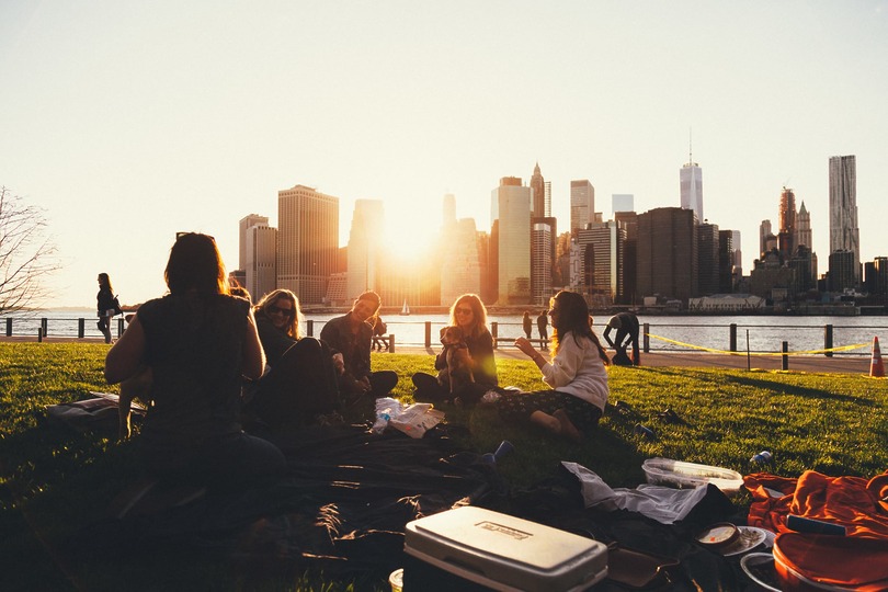 Group of people having a picnic in a park with a city skyline and sunset in the background