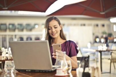 Woman smiling while looking at her phone, sitting at a desk with a laptop and glass under a red umbrella.