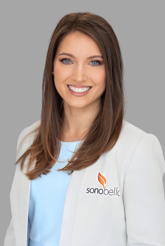Smiling woman with long brown hair wearing a white blazer with an orange logo and light blue shirt, gray background.