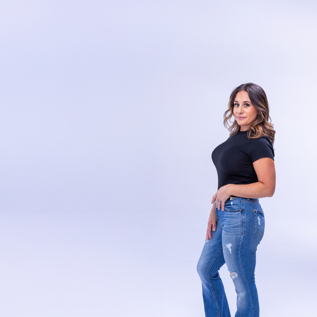 Woman in black top and blue jeans posing against a light purple background.
