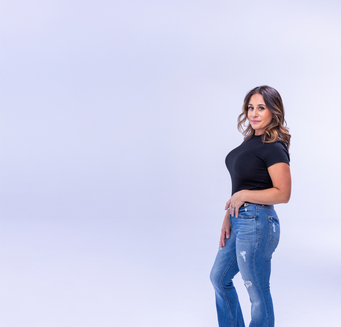 Woman in black top and blue jeans posing against a light purple background.