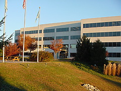 Modern office building with large windows surrounded by trees and grass on a sunny day, featuring flagpoles in the foreground.
