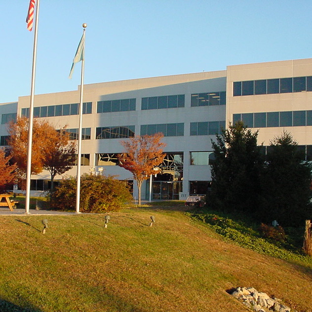 Modern office building with large windows surrounded by trees and grass on a sunny day, featuring flagpoles in the foreground.