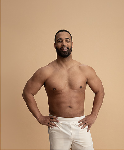 Man standing confidently with hands on hips, shirtless, wearing white pants, smiling against a beige background.
