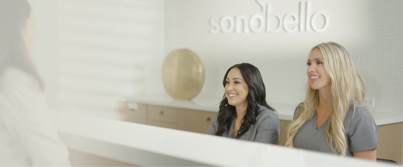 Smiling woman sitting at a reception desk with a company logo on the wall behind her.
