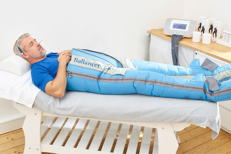 Man lying on a therapy bed wearing recovery compression sleeves, with medical equipment in the background.