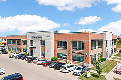Modern two-story office building with brick and white facade, surrounded by a parking lot with several cars on a clear day.