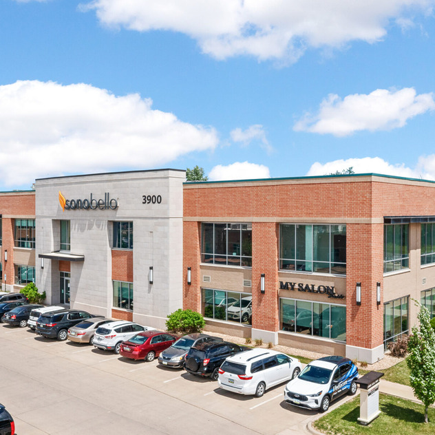 Modern two-story office building with brick and white facade, surrounded by a parking lot with several cars on a clear day.