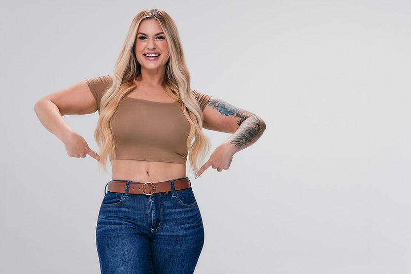 Sono Bello ambassador in jeans and a tshirt gestures toward flat belly after procedure