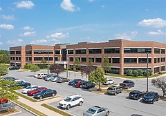 Office building with a brick facade, surrounded by a parking lot filled with cars under a clear blue sky.