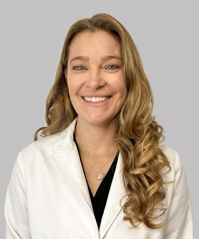 Smiling person with long wavy hair wearing a white coat against a plain background.