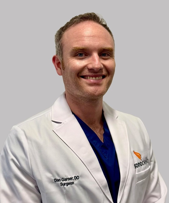 Man in a white medical coat with blue scrubs smiling against a gray background.