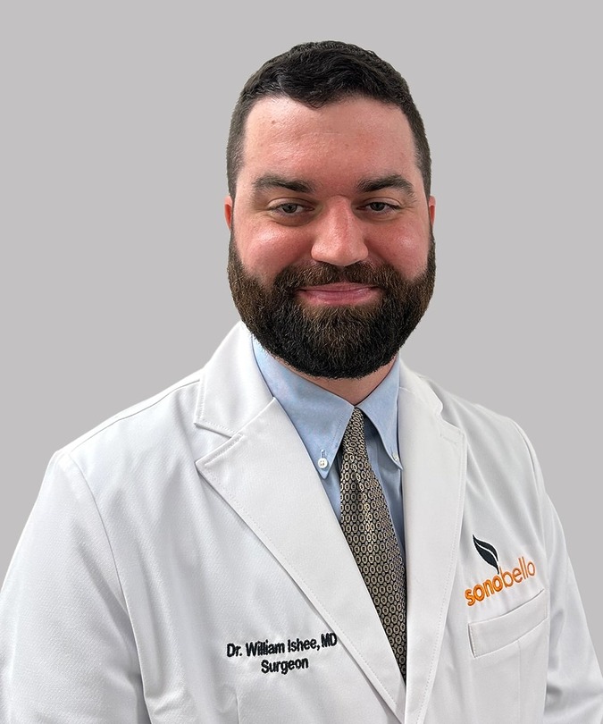 Male surgeon in a white coat with a beard and a light blue shirt, smiling against a plain gray background.