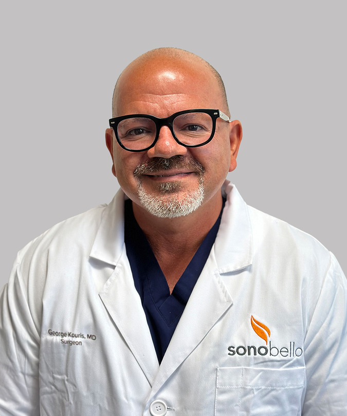 Man in glasses wearing a white medical coat with a logo, smiling against a gray background.