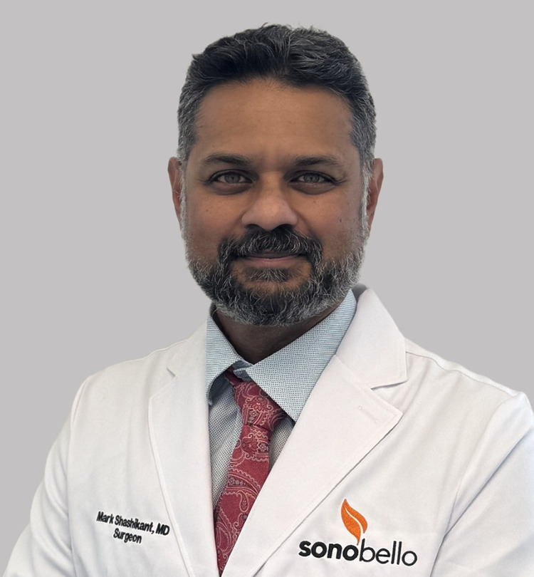 Male doctor in a white coat with a grey beard and a red tie, posing against a plain grey background.