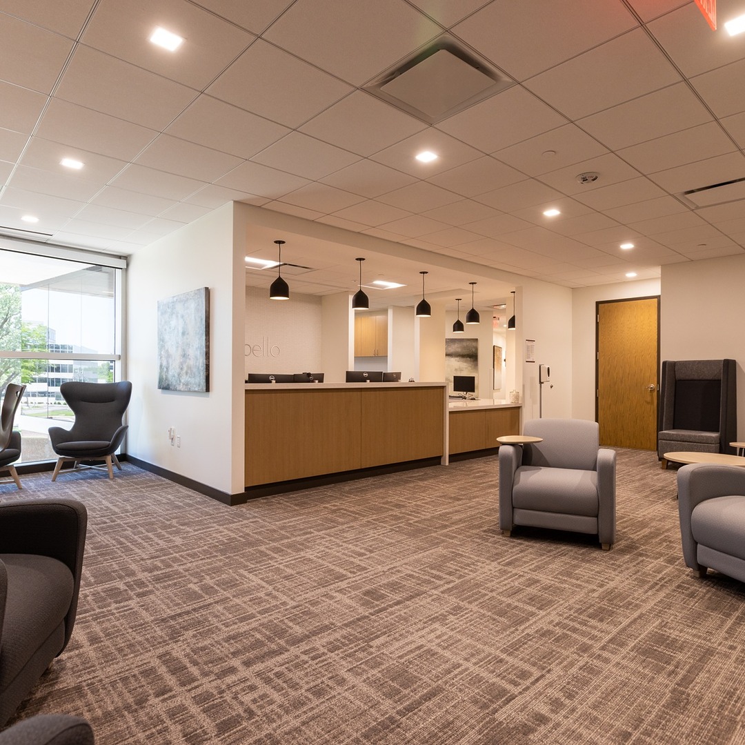 Modern office lobby with armchairs, ceiling lights, and a reception desk, featuring neutral tones and large windows.