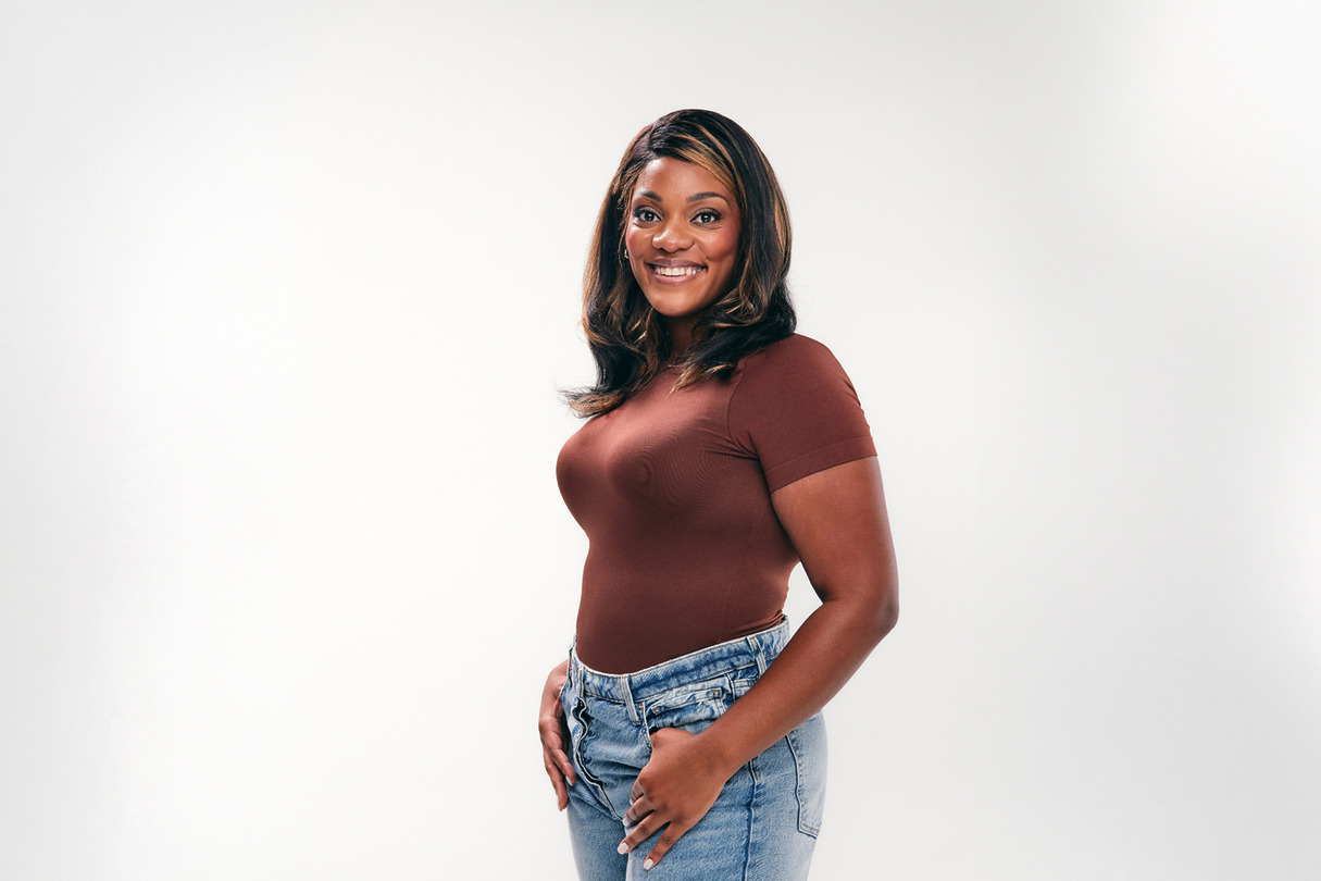 Smiling person in a brown top and blue jeans against a plain white background.