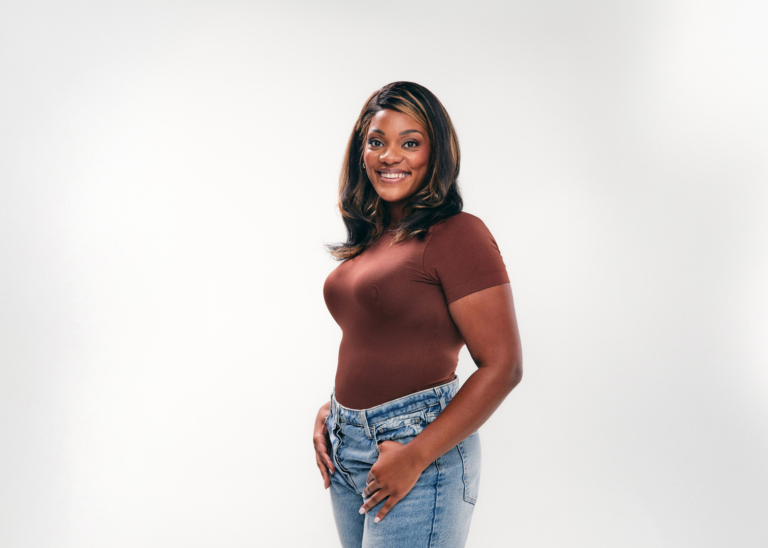Smiling person in a brown top and blue jeans against a plain white background.