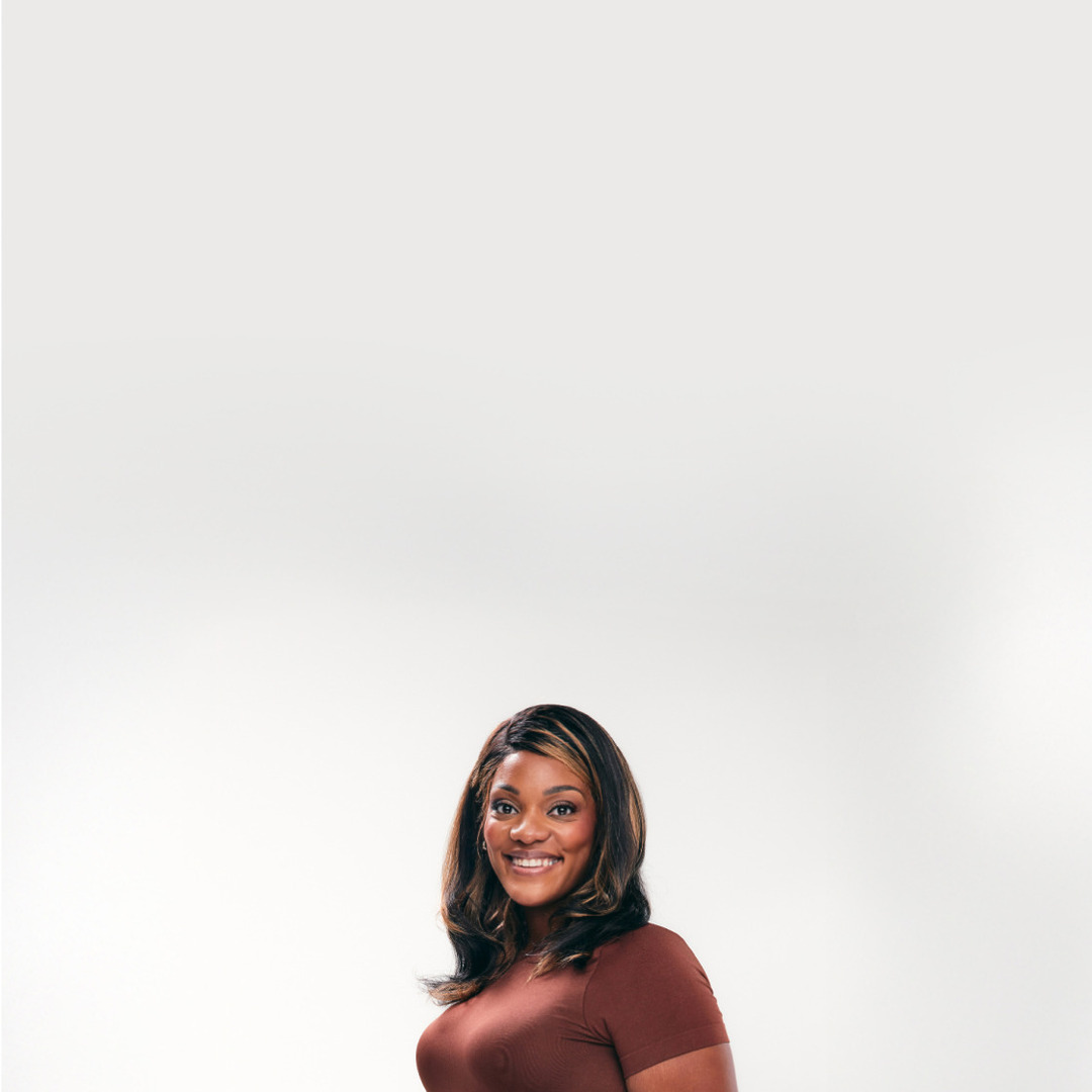 Smiling woman in a brown shirt and denim jeans standing against a plain white background.