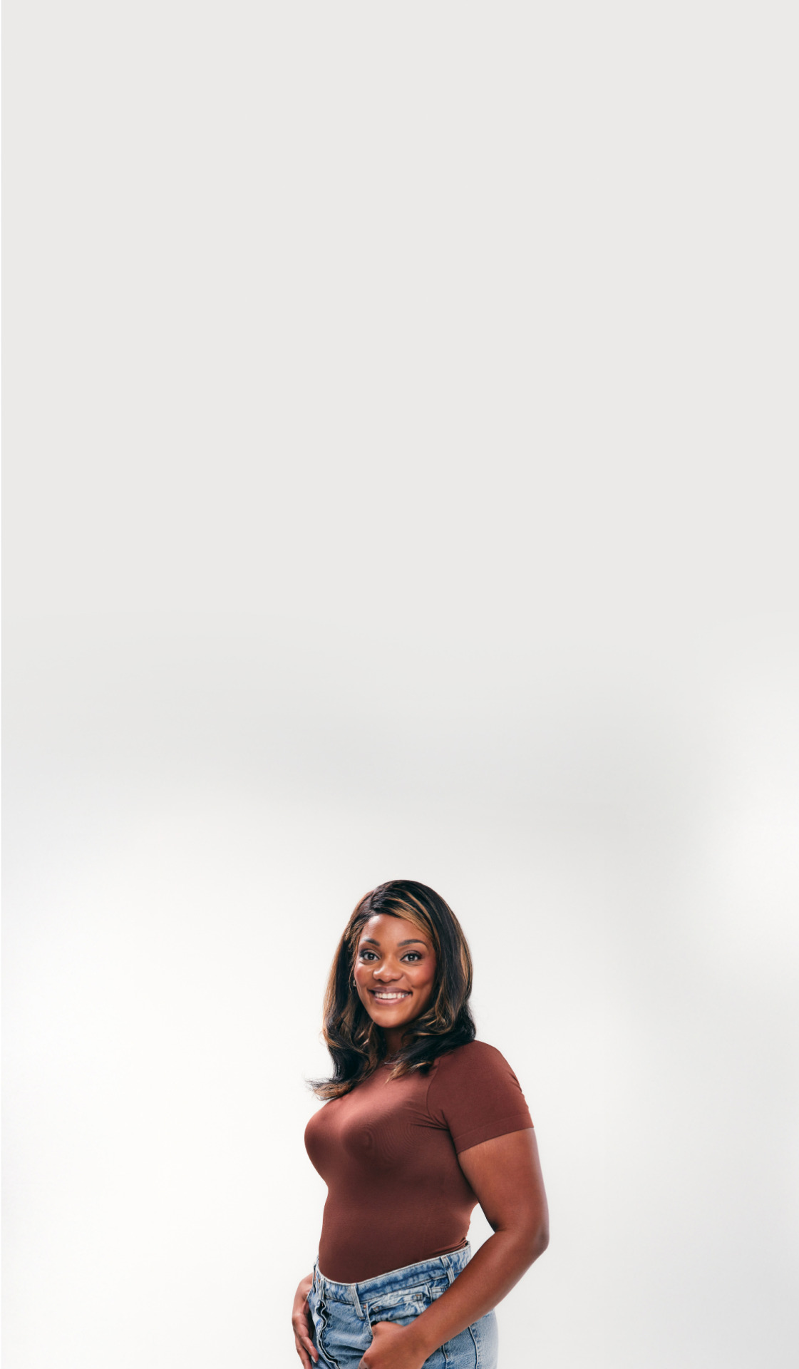Smiling woman in a brown shirt and denim jeans standing against a plain white background.