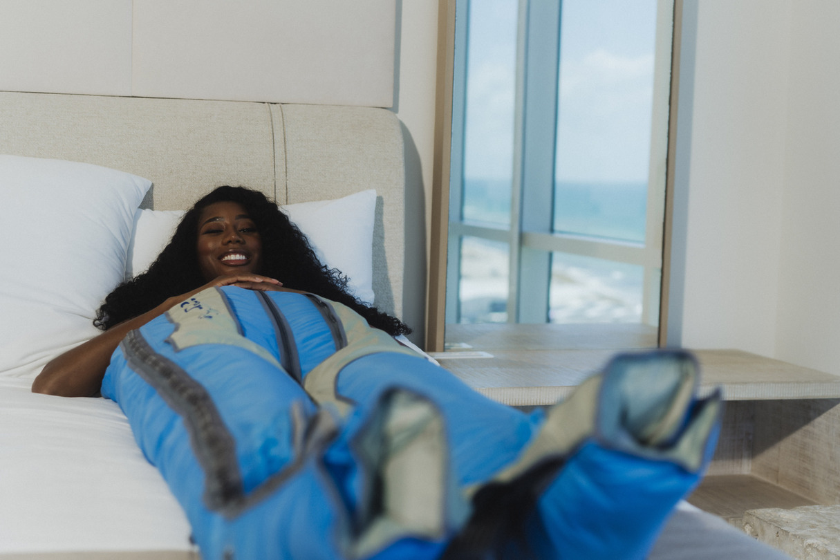 Woman relaxing on a bed, wearing a blue ballancer pro suit, beside a window with an ocean view in the background.