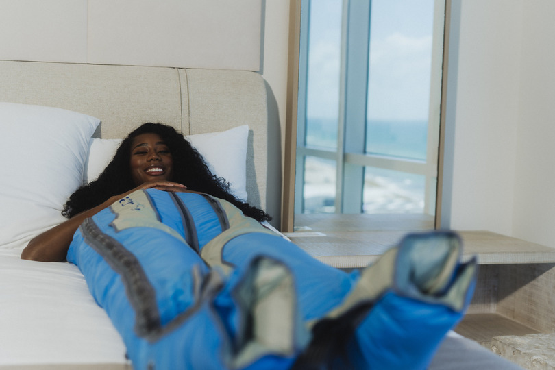 Woman relaxing on a bed, wearing a blue compression suit, beside a window with an ocean view in the background.
