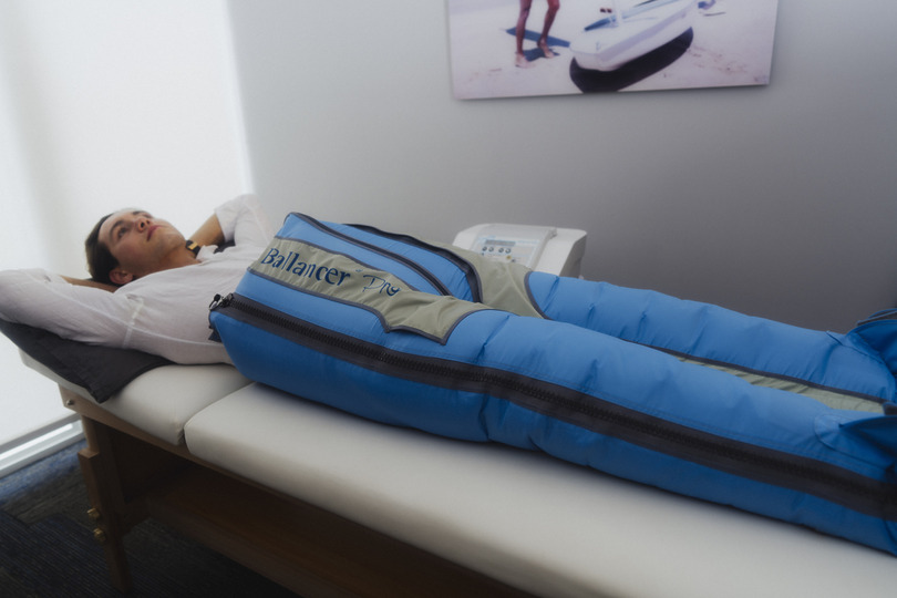 Person reclining on a therapy table, legs covered in a blue compression therapy device, relaxing in a wellness setting.