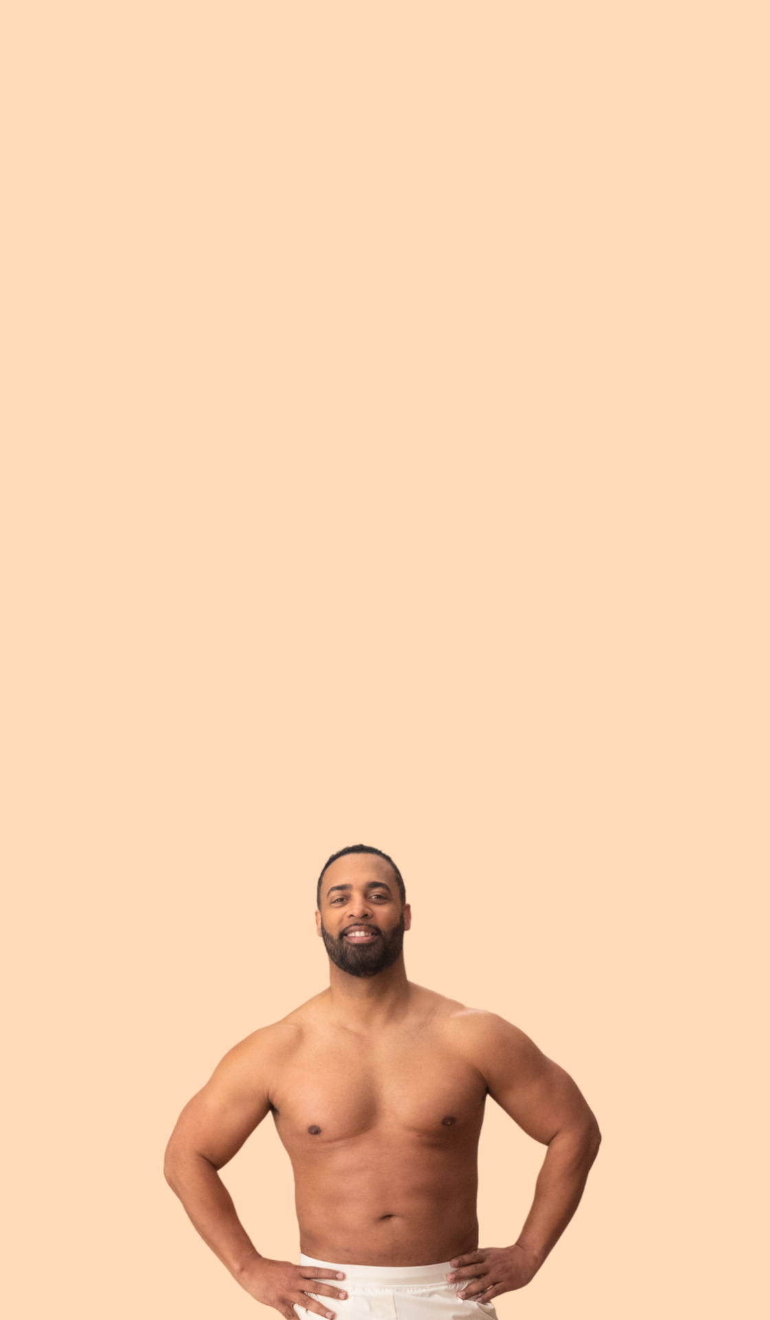Man with beard standing against a peach background, shirtless and wearing white pants, arms akimbo with a confident expression.