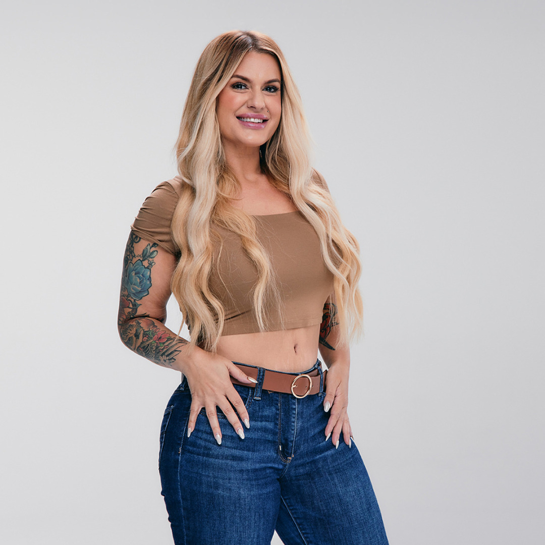 Woman with long blonde hair, wearing a brown crop top and blue jeans, posing confidently against a neutral background.