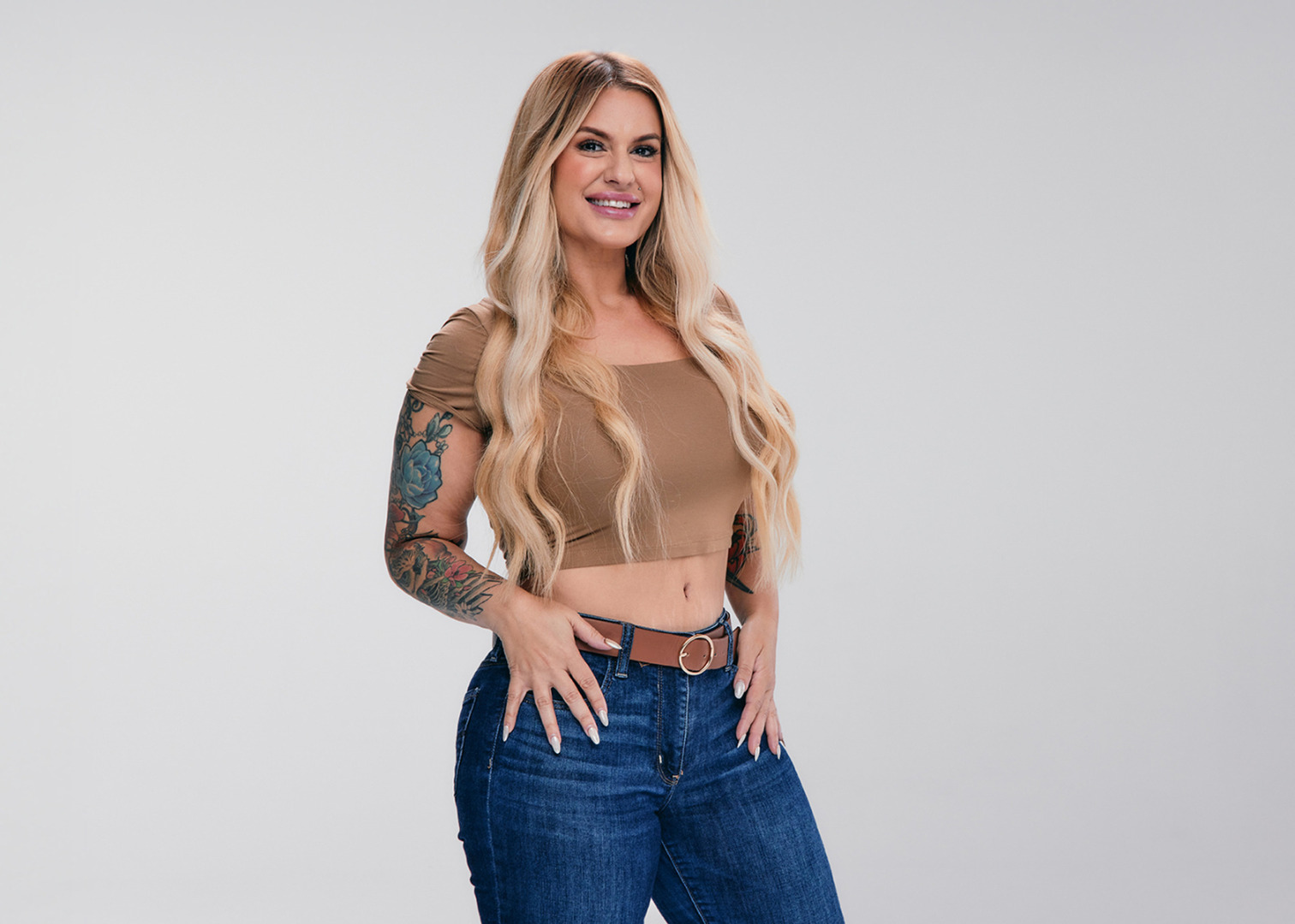 Woman with long blonde hair, wearing a brown crop top and blue jeans, posing confidently against a neutral background.
