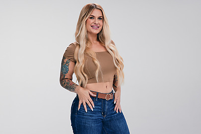 Woman with long blonde hair, wearing a brown crop top and blue jeans, posing confidently against a neutral background.