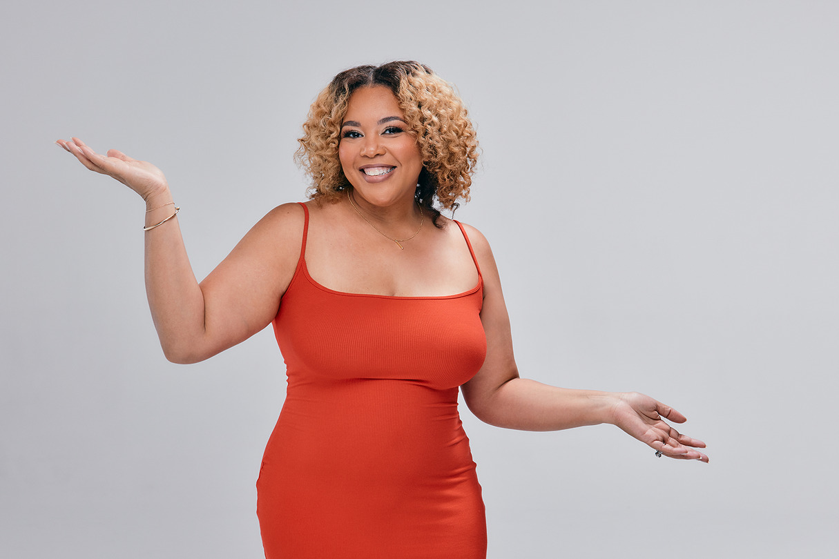 Person in a red dress smiling with arms raised against a gray background.