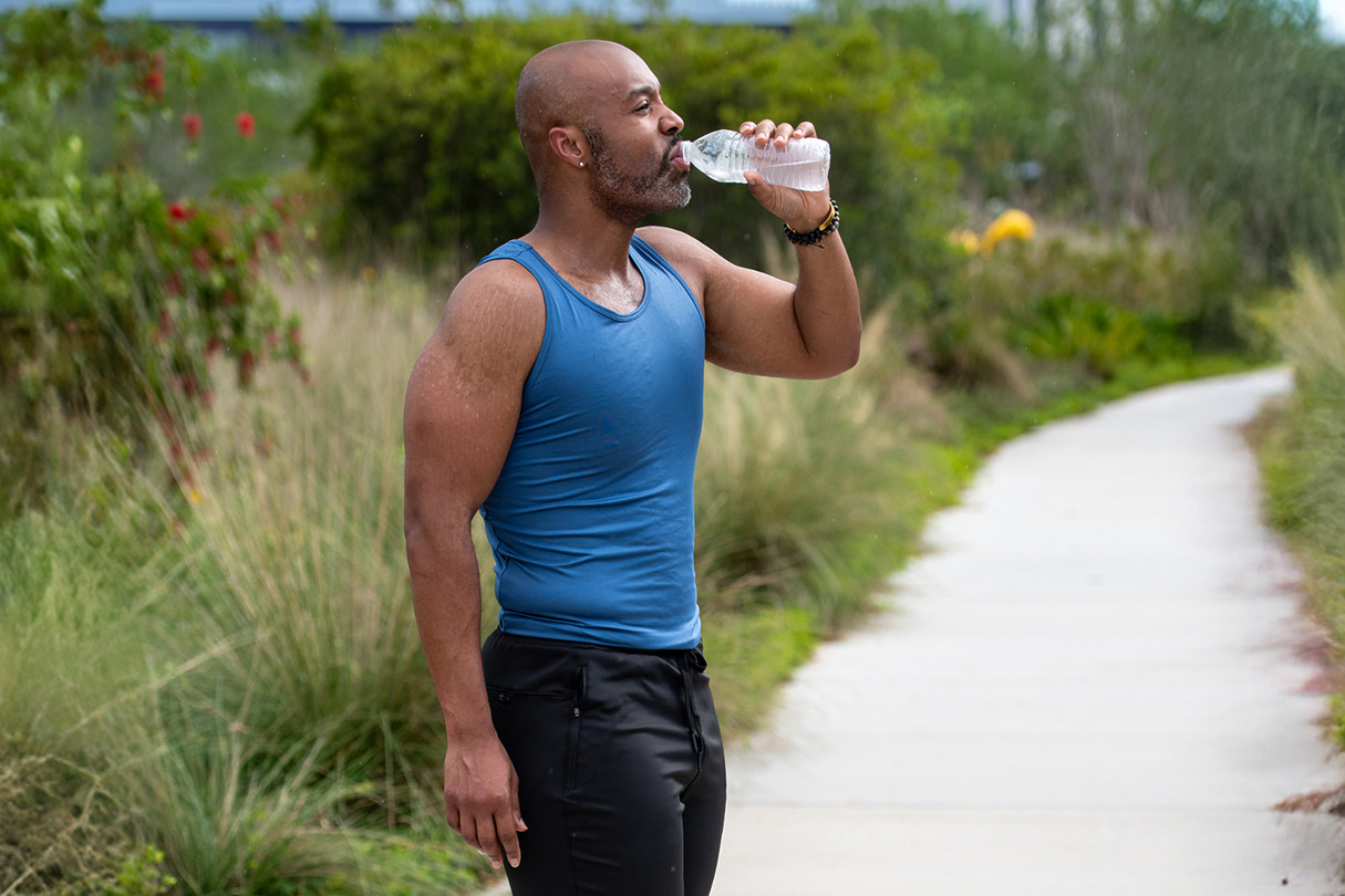 Man in a blue tank top drinks water from a bottle while standing on a pathway surrounded by greenery.