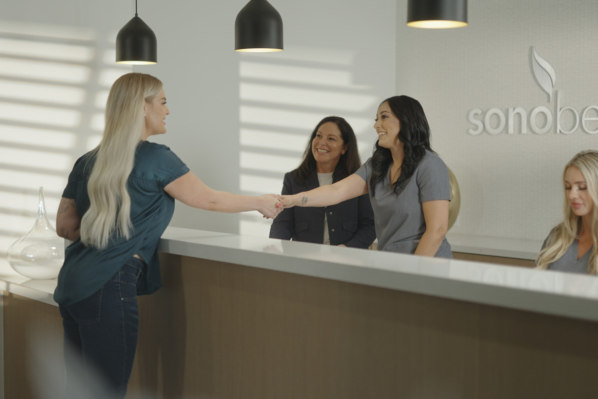 A woman shaking hands with a receptionist at a front desk in a modern office while two colleagues observe.