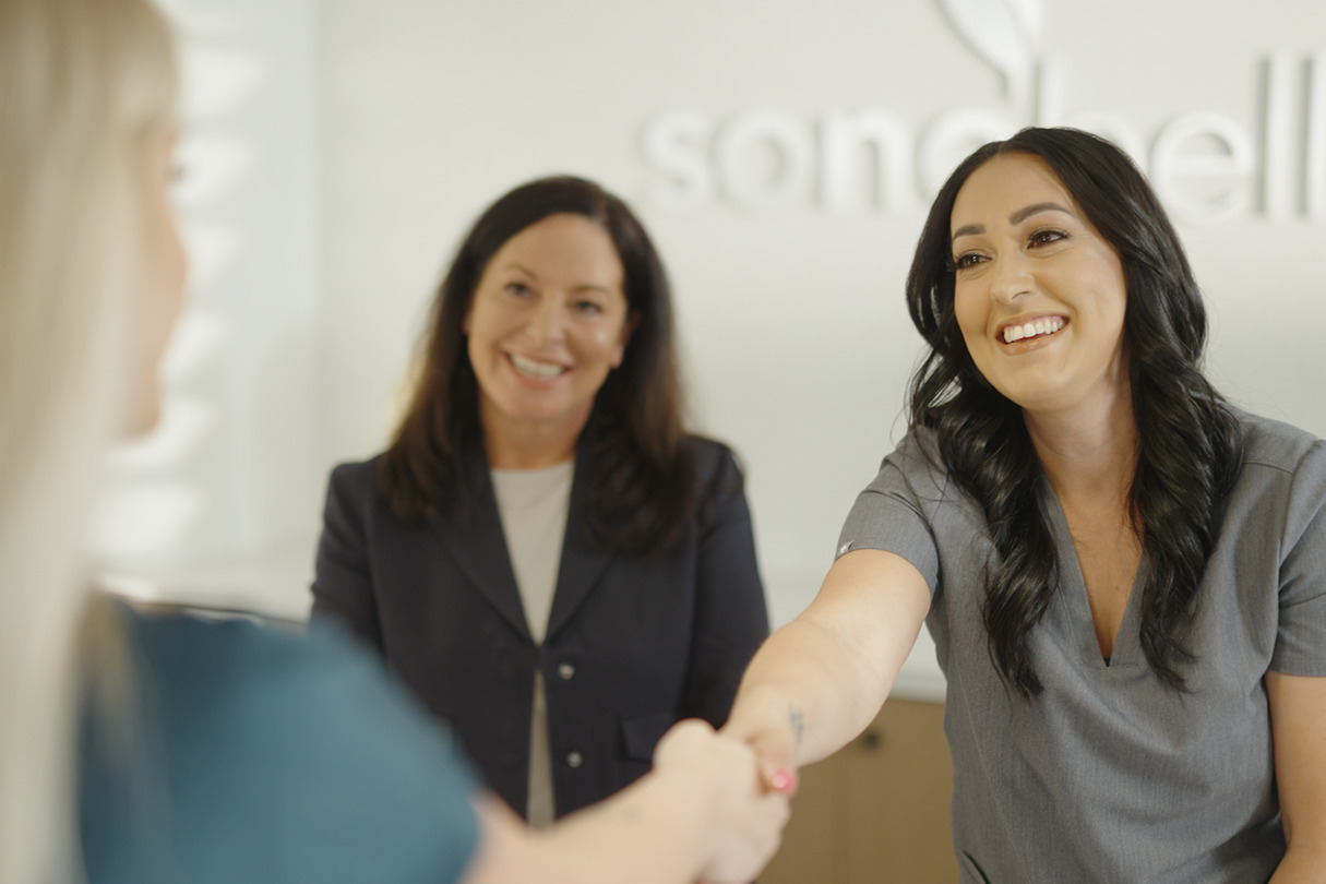Two women smiling and welcoming a new team member with a handshake in an office setting.