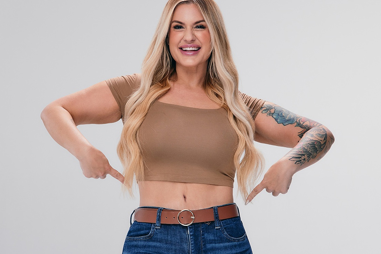 Smiling woman with long hair points to belt, wearing tan crop top and jeans, showcasing tattoos on her right arm.