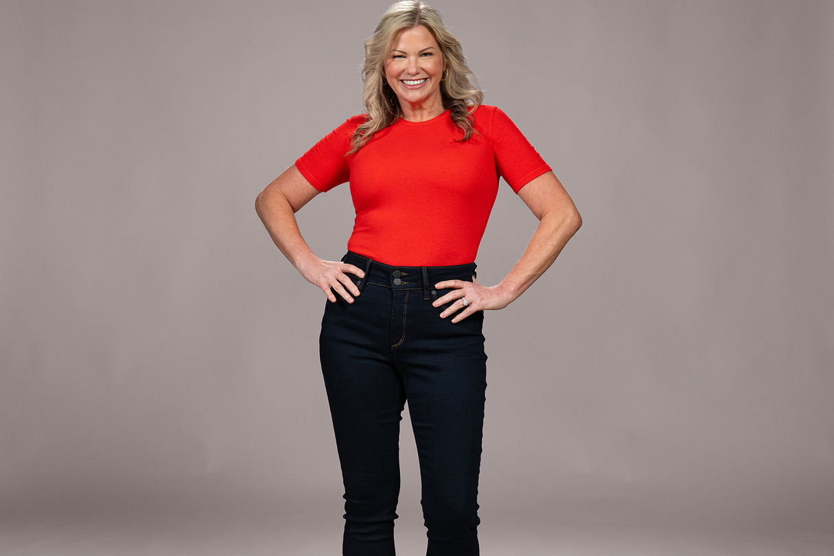 Person wearing a red shirt and dark jeans, smiling with hands on hips against a gray background.