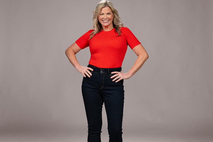 Person wearing a red shirt and dark jeans, smiling with hands on hips against a gray background.
