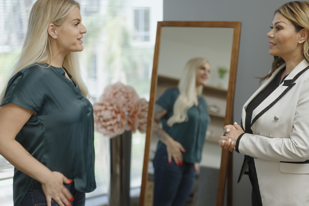 Two women having a conversation in front of a mirror, with one woman reflecting in the mirror.