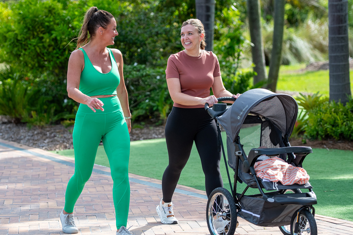 Two women walking and chatting in a park, one pushing a stroller, surrounded by greenery and sunlight.