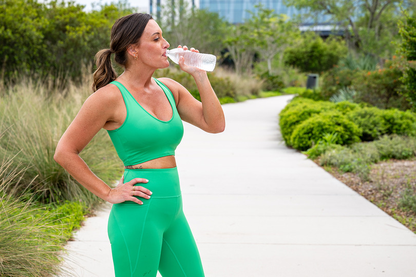 Woman in green activewear drinking water while standing on a park path with lush greenery around.